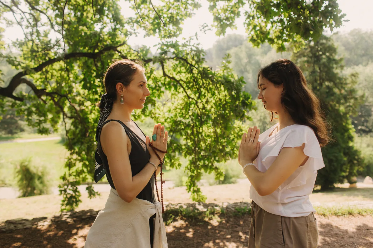 Zwei Frauen stehen sich unter einem Baum gegenüber, die Hände vor der Brust