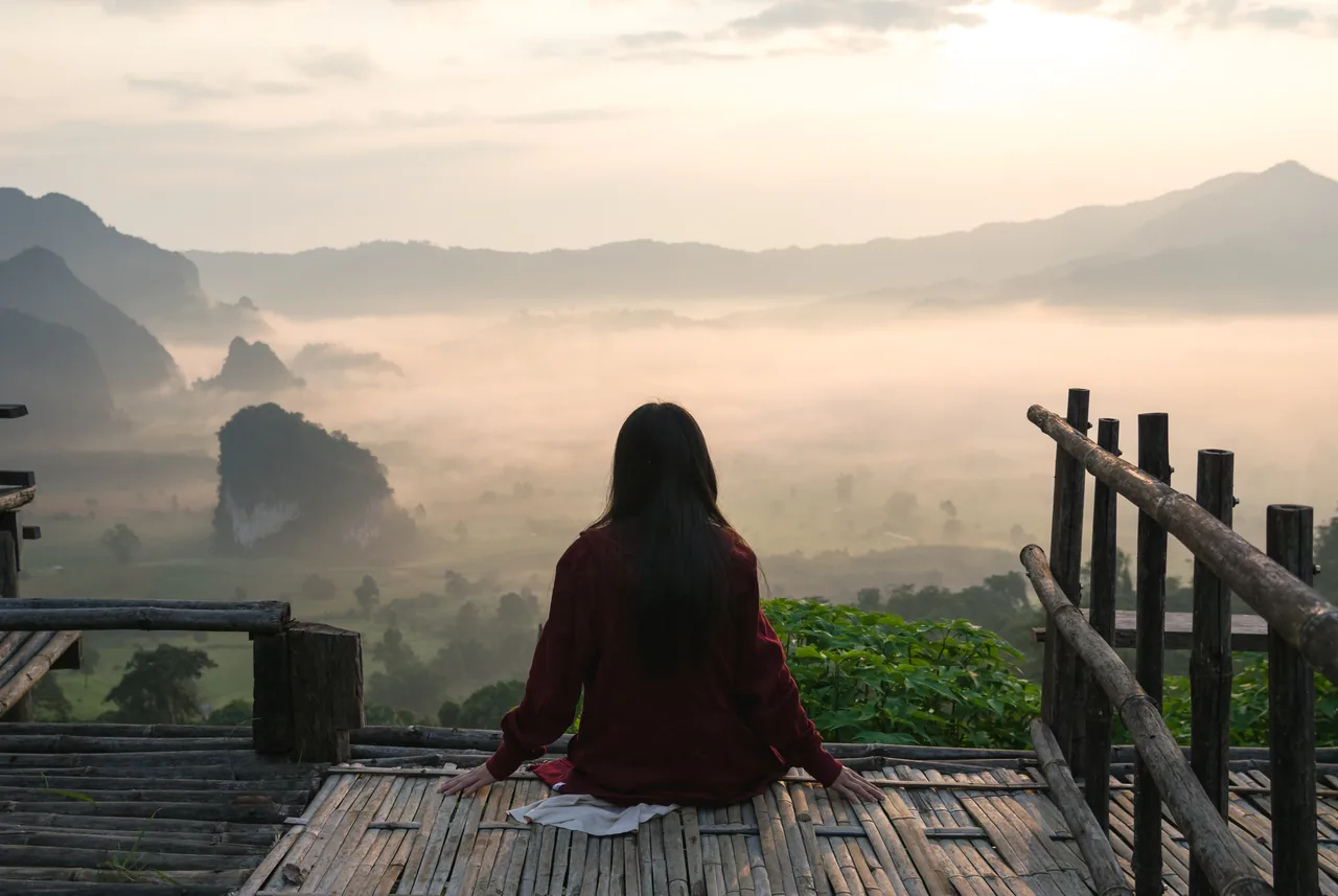 Eine Person sitzt allein auf einem Holzsteg vor einer Berglandschaft im Morgennebel